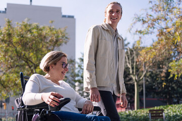 portrait of a woman using electric wheelchair with a young man talking happy as they walk through...