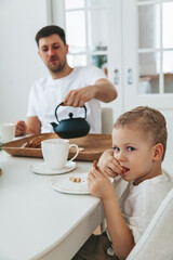 Father and his little son in white clothes drinking tea and eating a cake for the breakfast