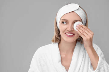 Young woman cleaning face with cotton pad on light grey background. Space for text