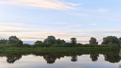 Fototapeta na wymiar On a quiet summer evening, after sunset, the sky with feathery clouds over the river turns pink and is reflected in the calm water. Shrubs and trees grow on the far grassy bank