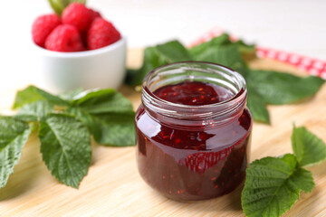 Jar of delicious raspberry jam and green leaves on wooden board, closeup. Space for text