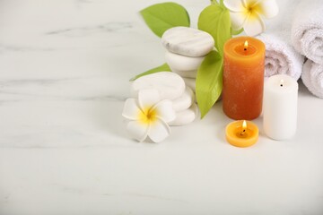 Fototapeta na wymiar Spa composition. Burning candles, plumeria flowers, stones and green leaves on white marble table, space for text