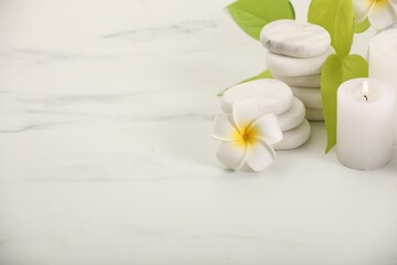 Spa composition. Burning candle, plumeria flower, stones and green leaves on white marble table, space for text