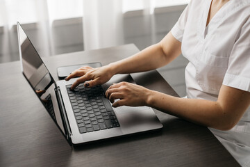 Close-up of a female family doctor working on a laptop computer in a clinic. A doctor in a white lab coat reviews a medical history at a table in a hospital office.