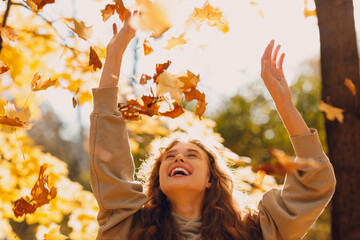 Smiling young woman with hands up throws up the autumn leaves in the forest at sunset