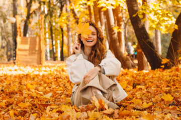 Smiling young woman sits in the autumn foliage on the ground of the forest with the yellow leaves
