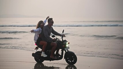 Romantic couple ride motorbike on ocean beach splashing water on a date at sunset. Lovers travel by motorcycle on coastline with reflection, splashes. Girlfriend hugs her boyfriend, spreads arms.