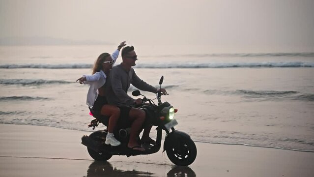 Romantic couple ride motorbike on ocean beach splashing water on a date at sunset. Lovers travel by motorcycle on coastline with reflection, splashes. Girlfriend hugs her boyfriend, spreads arms. - Powered by Adobe