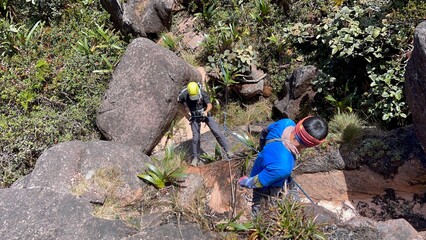 Hiker in safety harness and helmet climbing down a rope from rocky cliff under guidance of...