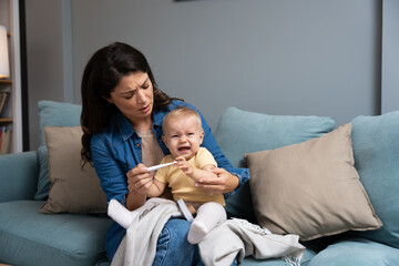Mother measuring temperature of her sick crying baby sitting on sofa in living room, little infant child catching cold, having influenza or grippe. Young mom worry for her kid cry - obrazy, fototapety, plakaty