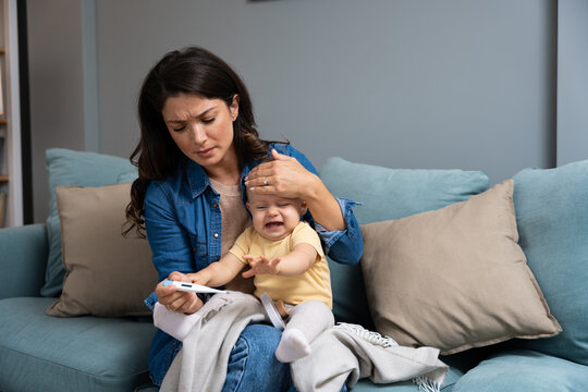 Mother measuring temperature of her sick crying baby sitting on sofa in living room, little infant child catching cold, having influenza or grippe. Young mom worry for her kid cry