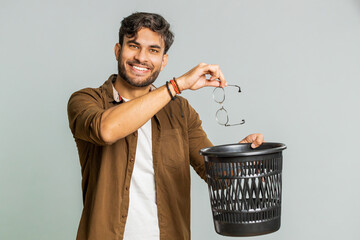 Indian young man taking off throwing out glasses into bin after medical vision laser treatment therapy surgery looking smiling at camera, heal, cure. Arabian guy isolated on studio gray background