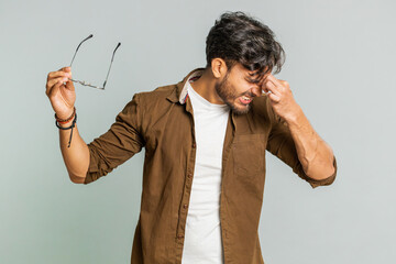 Exhausted tired Indian young man takes off glasses, feels eyes pain, being overwork burnout from long hours working. Sleepy exhausted Arabian guy rubbing eyes isolated on studio gray background - obrazy, fototapety, plakaty