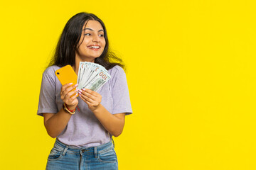Indian woman looking smartphone display sincerely rejoicing win, receiving money dollar cash banknotes celebrating success victory play lottery game. Lucky rich Arabian girl on yellow background - obrazy, fototapety, plakaty