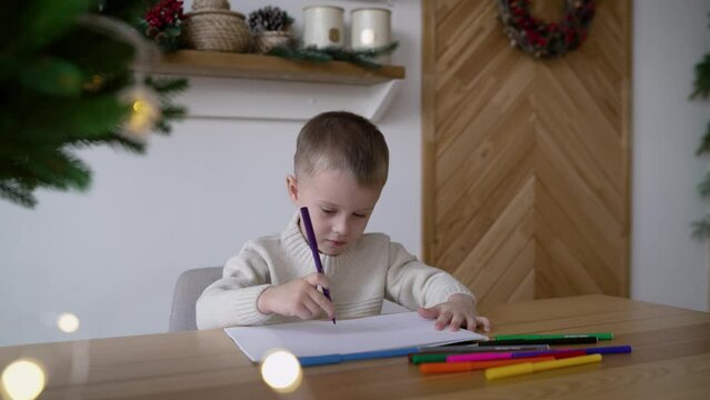 A boy writes wishes to Santa Claus. A little boy, sitting at the table, draws a New Year's interior. A boy draws while sitting at a table.