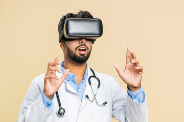 Excited happy Indian doctor cardiologist man using headset helmet app, watching virtual reality 3D 360 video. Arabian apothecary guy in VR goggles on beige background. Future medicine technology