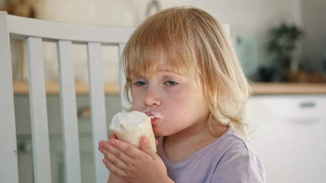 Baby girl enjoying ice cream. Pretty little toddler eating an ice-cream indoors, at home. Dining room background. Small child eats plombir and cream messy on her mouth. Cute kid with tasty sweet food.