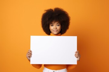 young woman holding a blank white poster or sign over a orange studio background with copyspace