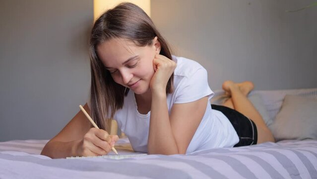 Cheerful positive smiling brown haired woman dressed in pajama makes notes in diary lies on bed writing letter to her boyfriend enjoying her text having dreaming expression.