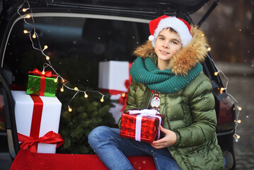 child boy in a green knitted scarf and a Santa hat sits in the trunk of a car with a gift in his hands