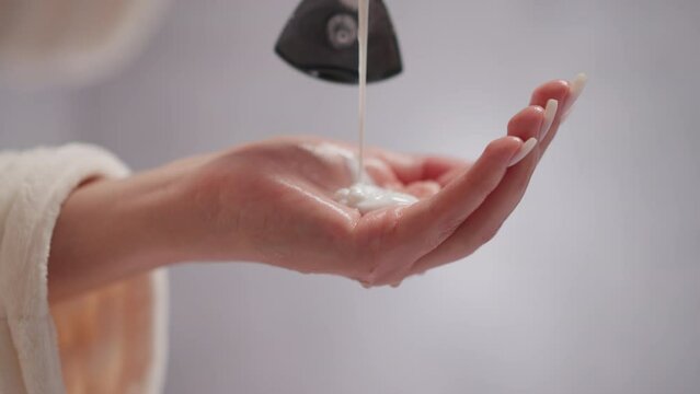 Person pours liquid conditioner for hair into palm in shower. Creamy hair conditioner drips onto female hand in spacious room with blurred background