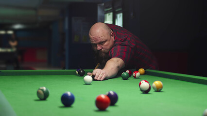 A Big Bald Brutal Bearded Man Plays Billiards On A Green Table, Takes Aim And