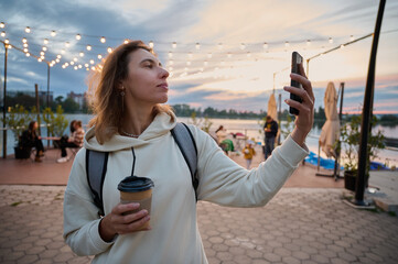 Portrait of pretty woman walking on the beach, drinking tea, taking a selfie and browsing social networks on the phone. Beautiful sunset on the beach and beautiful artificial lighting in a beach cafe