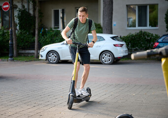 Young sporty man in a striped T-shirt riding an electric scooter in the park. Rent an electric...