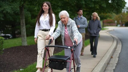 Grandmother using a walker and granddaughter smiling and walking together outside with rest of family following behind.
