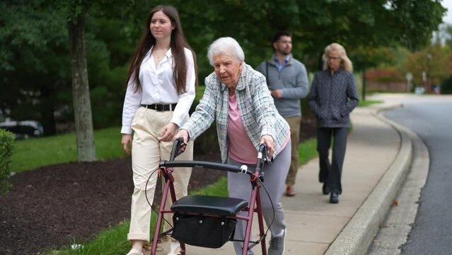 Grandmother using a walker and granddaughter smiling and walking together outside with rest of family following behind.