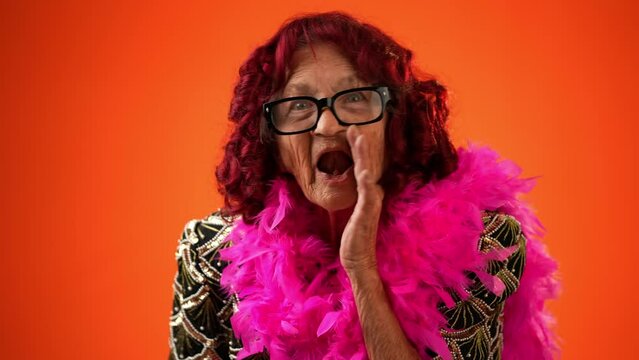 Funny portrait of smiling happy crazy toothless grandmother with wrinkled skin and boa puts hand to mouth to tell a secret isolated on orange background studio