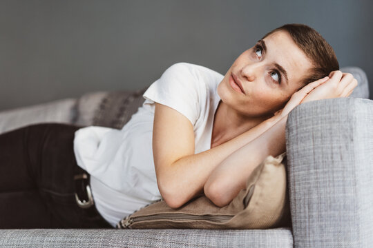 Young Modern Woman with Short Hair Relaxes on Sofa, Gazing Thoughtfully Upwards in Apartment