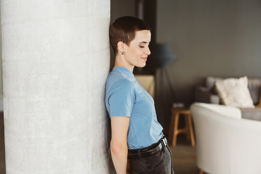 Young Modern Woman with Short Hair Leaning by a Pillar in an Apartment, Gazing Relaxedly Downward