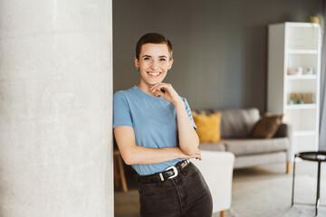 Confident Young Woman with Short Hair Leaning against Pillar in Apartment, Laughing at Camera - obrazy, fototapety, plakaty
