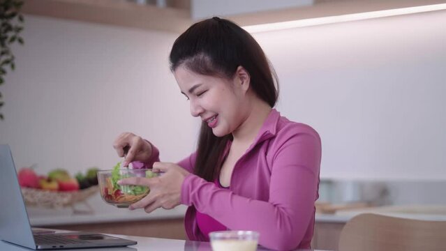 Sporty Young Asian Woman In Sportswear Looking At Laptop And Eating Vegetables Salad In The Kitchen Interior, Healthy Diet And Sport Concept.