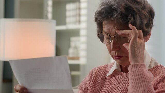 Old 60s Caucasian woman retired lady 70s senior aged female granny reading news open paper envelop read letter bad vision blurry eyesight correct glasses poor eyestrain check correspondence at home