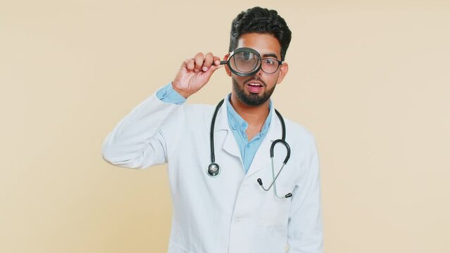 Investigator researcher scientist Indian young doctor cardiologist man holding magnifying glass near face looking , searching, analyzing. Arabian apothecary pharmacy guy isolated on beige background