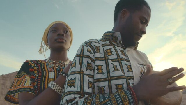 Chest up slowmo of African American man tapping on djembe drum while his beautiful wife standing behind him being alone together in desert