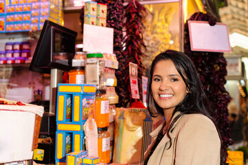 In a traditional market setting, a young and beautiful Hispanic woman smiles warmly, her gaze fixed directly on the camera