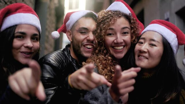 Diverse group of young people celebrating Christmas while singing and dancing together outdoors - Multiracial friends wearing Santa Claus hats on winter holidays
