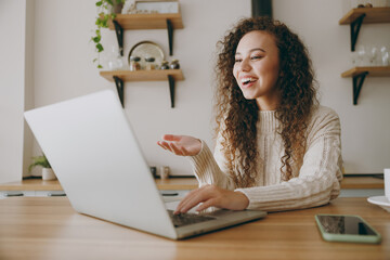 Young smiling housewife IT woman wear casual clothes sweater hold use work on laptop pc computer get video call talk speak sit at table in light kitchen at home alone. Lifestyle cooking food concept.