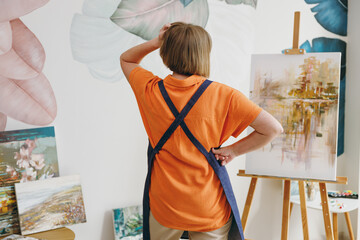 Back rear view of elderly caucasian european artist woman 50 years old wear casual clothes standing near easel with painting artwork spend free spare time in living room indoor. Leisure hobby concept - obrazy, fototapety, plakaty