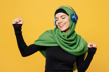 Young fun arabian asian muslim woman wearing green hijab abaya black clothes listen to music in headphones dance isolated on plain yellow background. People uae middle eastern islam religious concept.