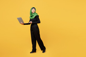Full body young arabian asian muslim IT woman wears green hijab abaya black clothes work hold use laptop pc computer look aside isolated on plain yellow background. People uae islam religious concept.
