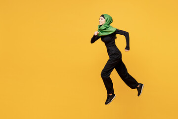 Full body side profile view young arabian asian muslim woman wear green hijab abaya black clothes jump high run fast hurry up isolated on plain yellow background. People uae islam religious concept.