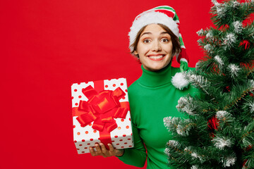 Young surprised fun woman wear green turtleneck Santa hat posing hold Christmas tree present box with gift ribbon bow isolated on plain red background. Happy New Year 2024 celebration holiday concept.