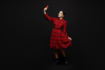 Full body young woman with Halloween makeup face art mask wear clown costume red dress do selfie shot on mobile cell phone isolated on plain solid black background studio. Scary holiday party concept.