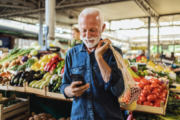 Mature cheerful man at the vegetable market shopping for vegetables and looking at the shopping list on his phone. Eldery man shopping on the market and using his smartphone.