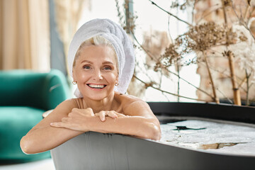 pleased middle aged woman with white towel on head taking bath in modern apartment, home spa