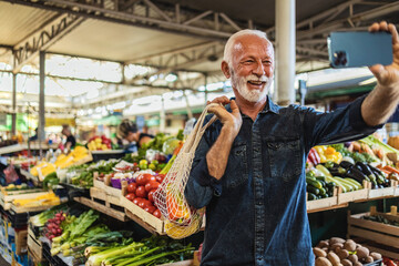 Joyful senior Caucasian man standing at  farmer's market using his mobile phone to take a selfie.
